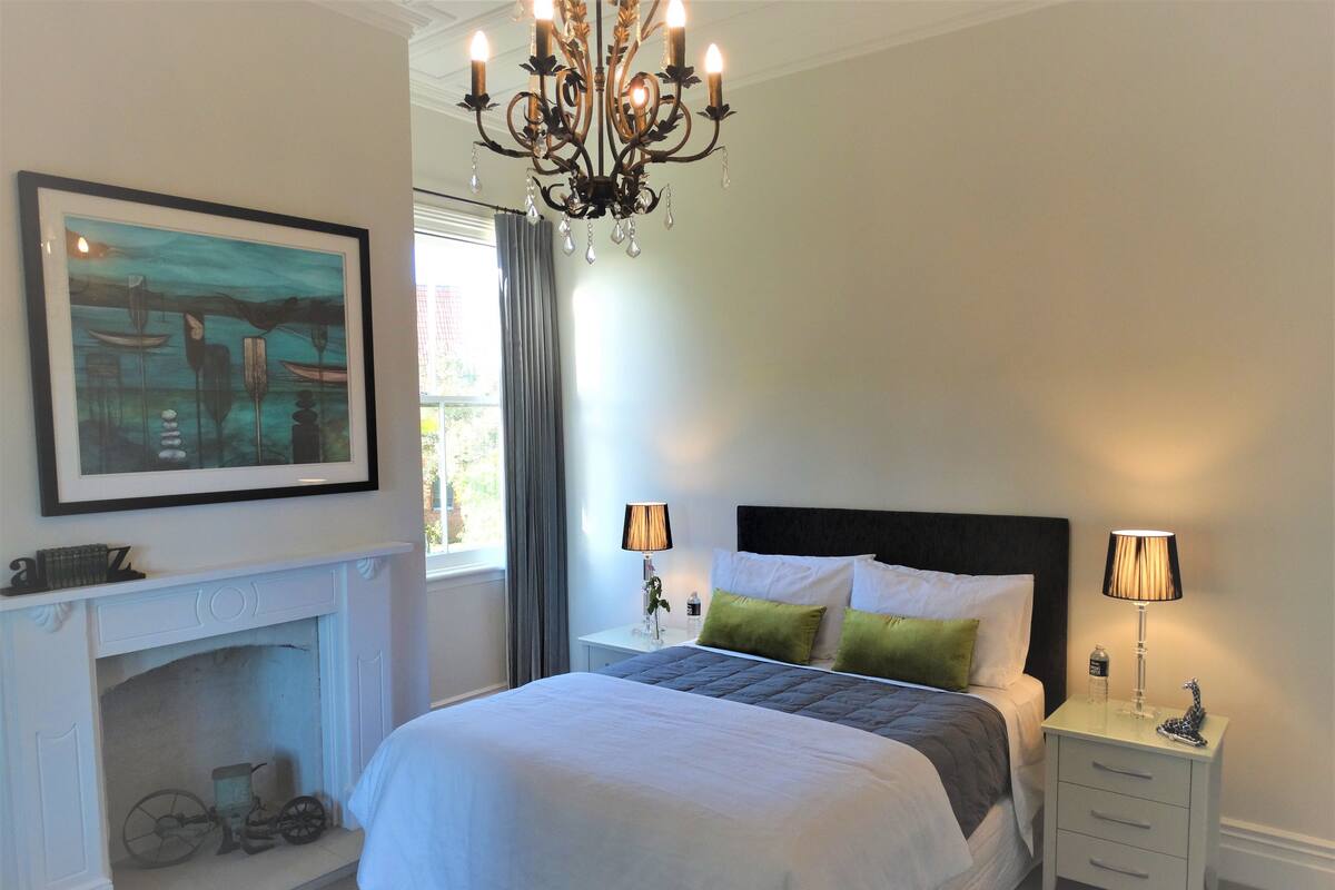 A bedroom with a large bed, two bedside lamps, a chandelier, a fireplace, and a framed picture on the wall.