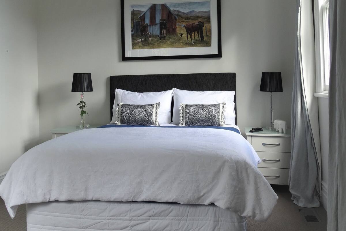 A neatly made bed with white linens, a black headboard, and a framed painting of a rural scene above it.