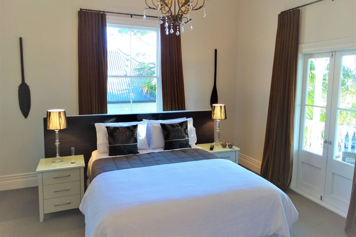 A bedroom with a large bed, two bedside tables, a chandelier, and a window with curtains.