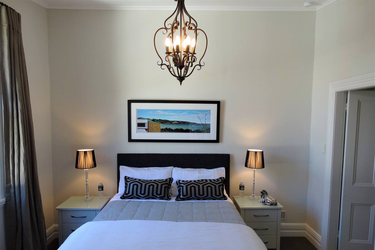 A neatly arranged bedroom with a large bed, two bedside tables with lamps, a chandelier, and a framed picture on the wall.