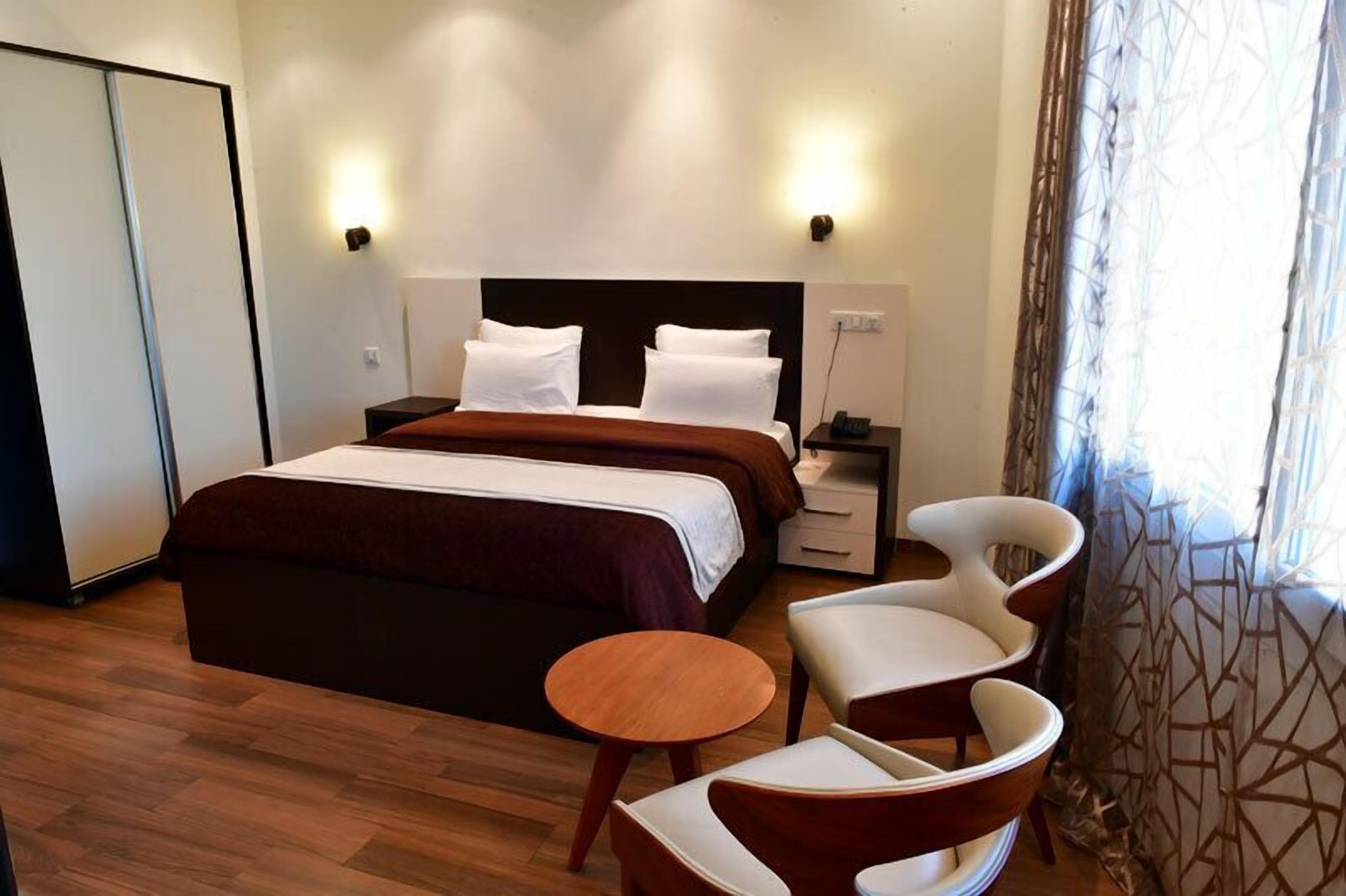 A hotel room with a bed, bedside tables, a chair, a small table, and a window with curtains.