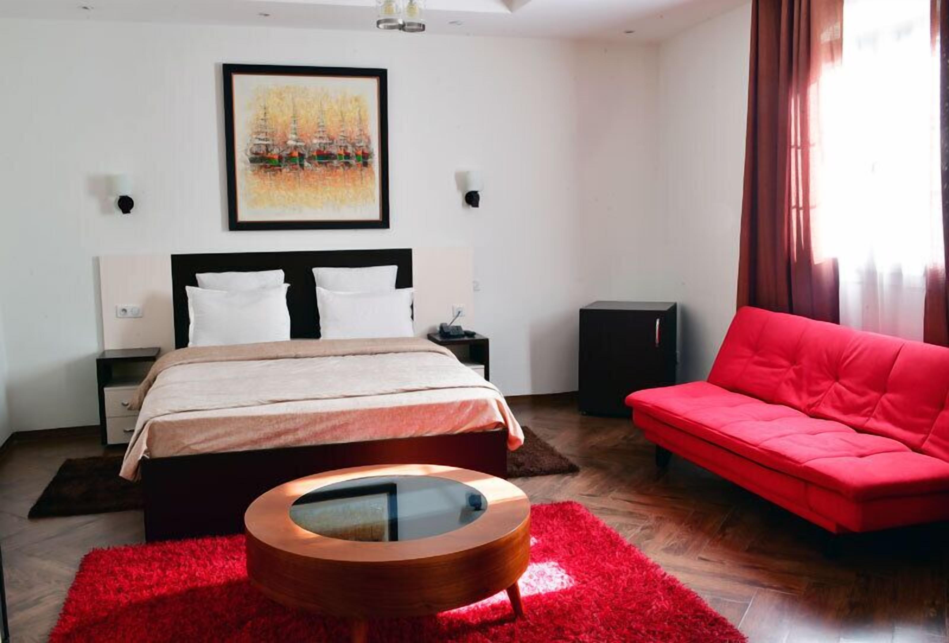 Un dormitorio con una cama, un sofá rojo, una mesa de centro de madera, un cuadro en la pared y un altavoz.