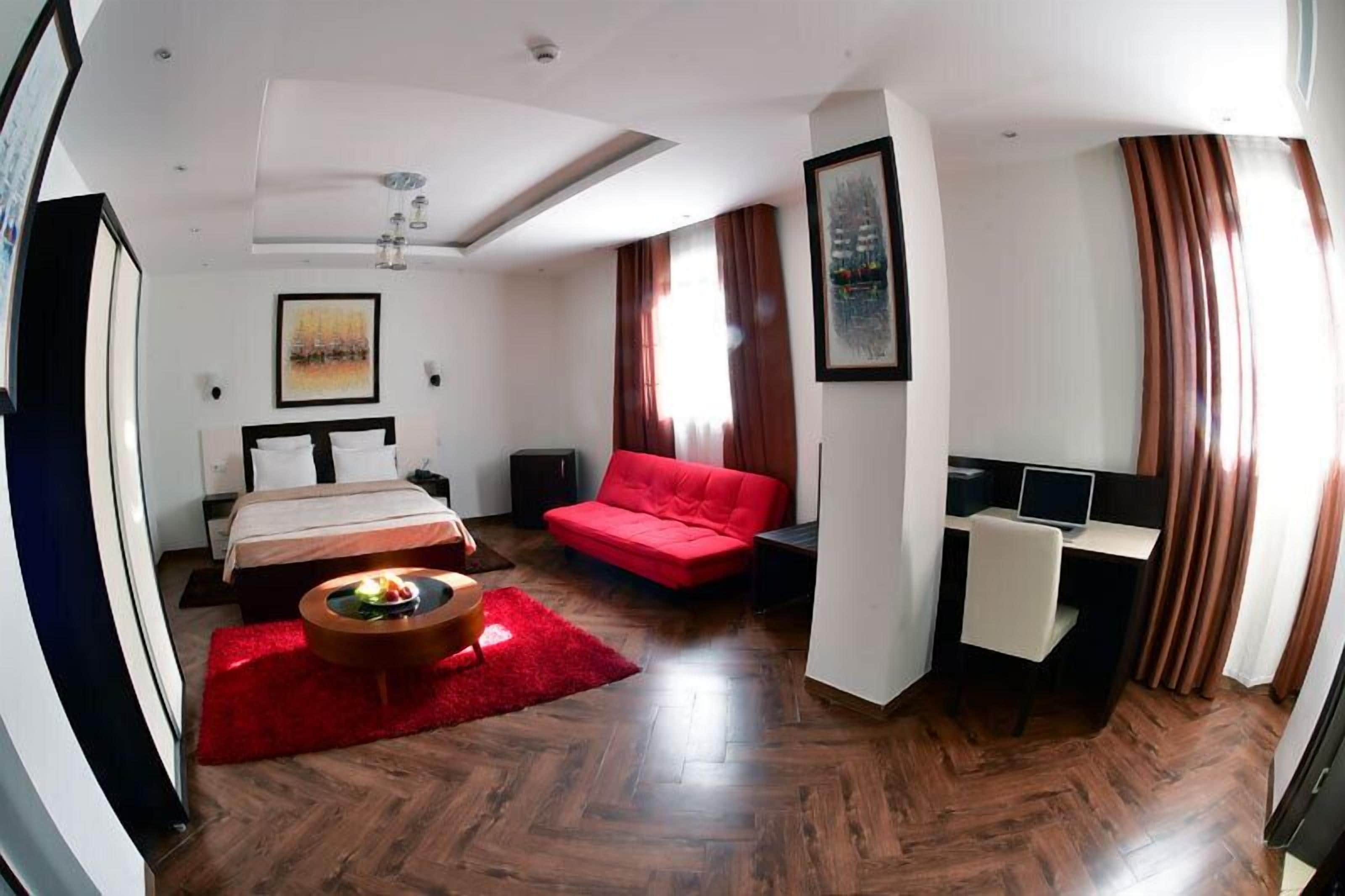 Una habitación de hotel moderna con una cama, un sofá rojo, un escritorio con computadora y una alfombra roja.