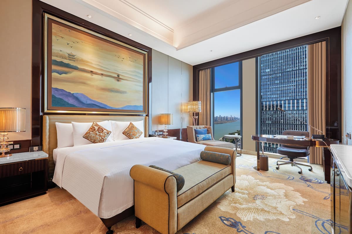 A hotel room with a large bed, a desk, a chair, and a view of the city through large windows.