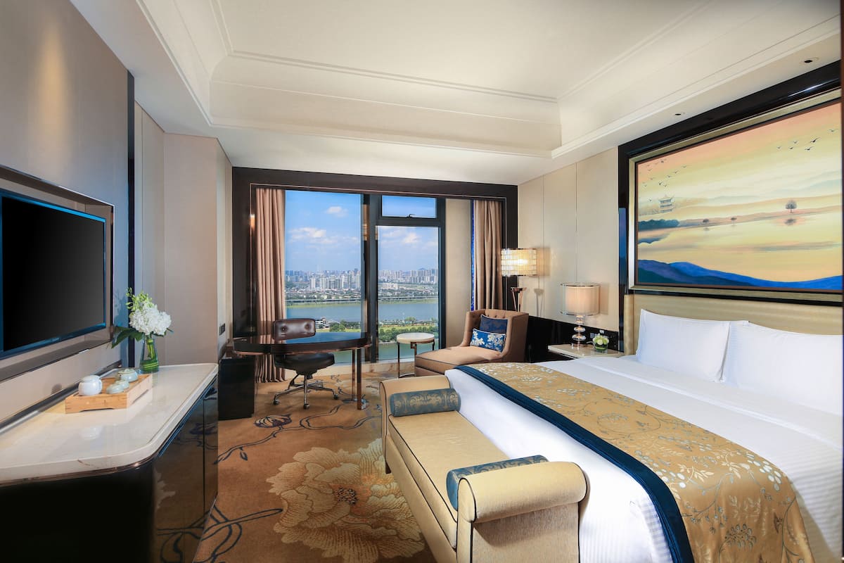 A hotel room with a large bed, a desk with a flat-screen TV, a chair, a small table, and a view of a cityscape through the window.