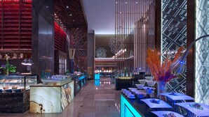 Un moderno vestíbulo de hotel con un mostrador de recepción de mármol, una vitrina con alimentos y una pared decorativa con diseños geométricos.