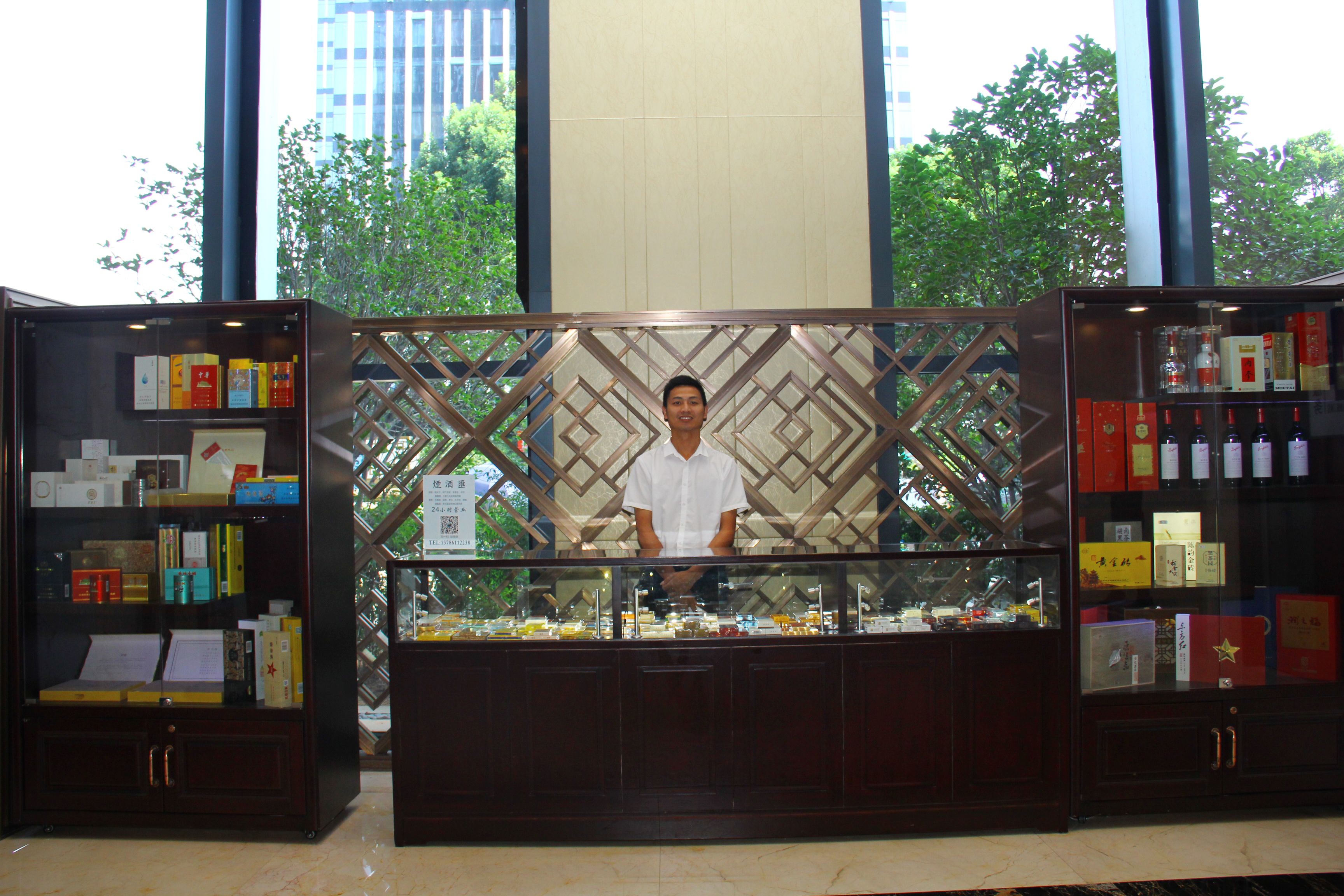 A man standing behind a display case with various items, in a room with large windows and a view of trees.
