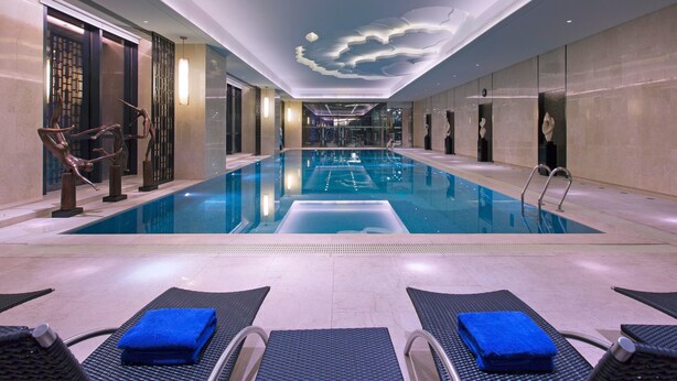 Una piscina interior moderna con agua cristalina, sillas de descanso con toallas azules y un techo decorativo con motivos de nubes.