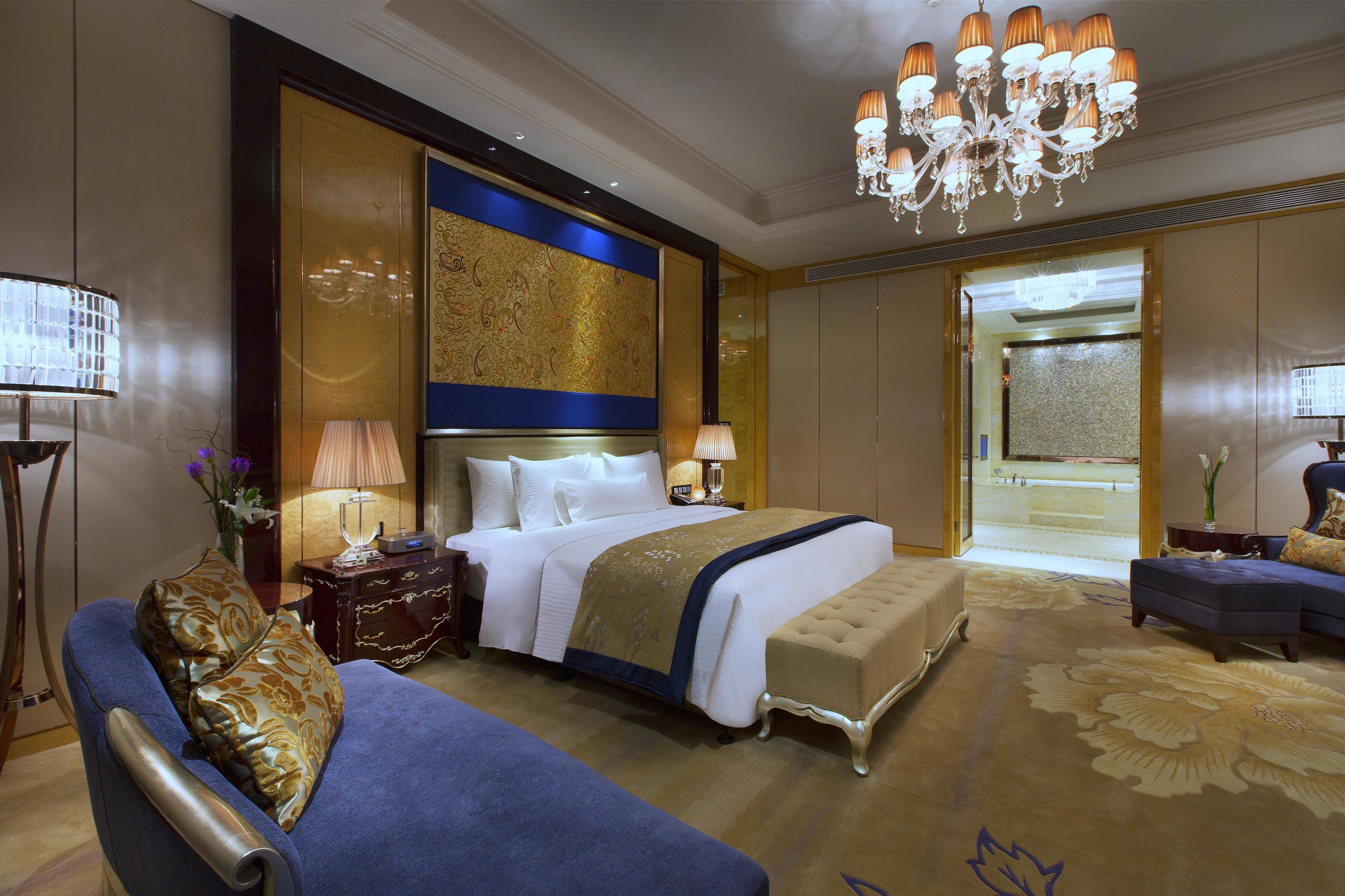 A spacious hotel room with a large bed, a seating area with a blue sofa, a chandelier, and a view of the lobby.