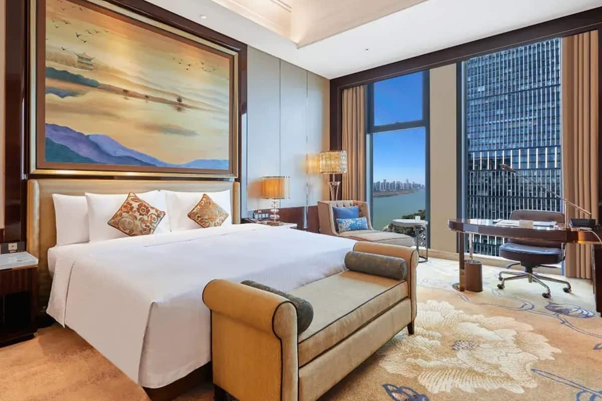 A hotel room with a large bed, a desk, a chair, a view of a cityscape, and a painting on the wall.