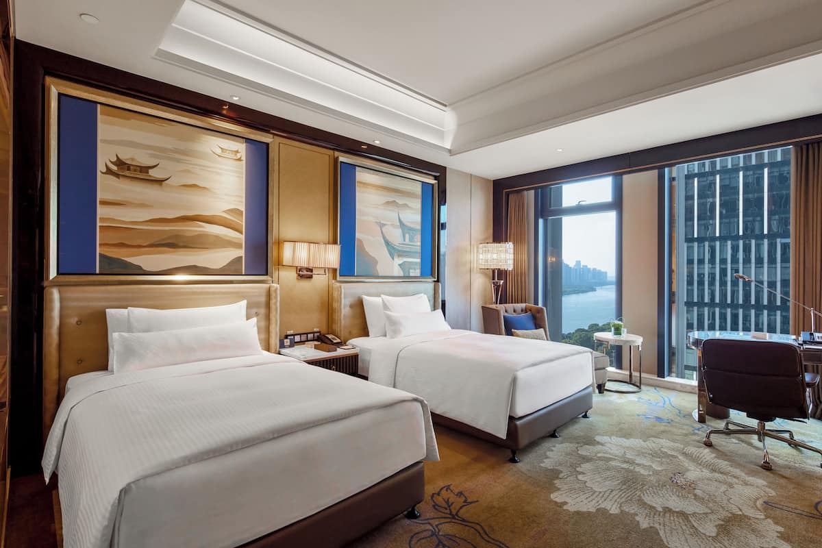 A hotel room with two beds, a desk, a chair, and large windows with a city view.