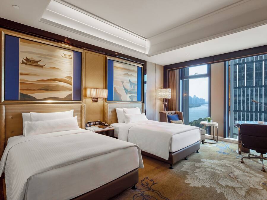 A hotel room with two beds, a desk, a chair, and large windows with a city view.