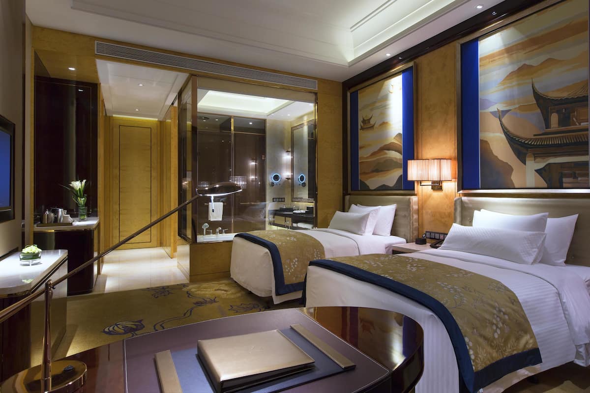 A hotel room with two beds, a desk with a lamp, a mirror, and a painting on the wall.