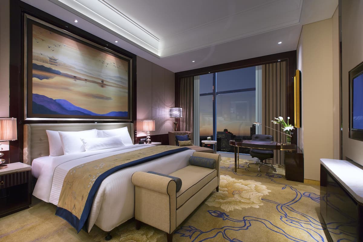 A hotel room with a large bed, a desk, a chair, a TV, and a painting on the wall.