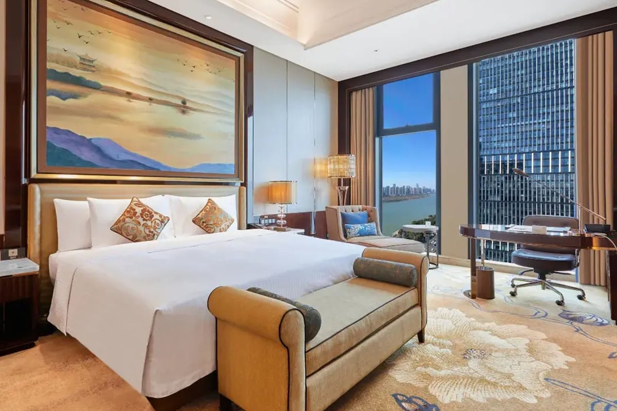 Une chambre d’hôtel avec un grand lit, un bureau, une chaise, une vue sur la ville et un tableau au mur.