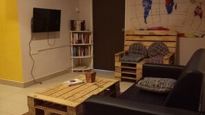 A cozy living area with a wooden coffee table, a black sofa, and two patterned armchairs. A bookshelf and a flat-screen TV are mounted on the wall. - HOOD - Hostel (Kota Kinabalu)