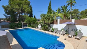 A rectangular swimming pool with a curved edge, surrounded by lounge chairs and umbrellas, with trees and a white wall in the background. - Casa Virginia 6 pax - Javea, Costa Blanca (Javea)