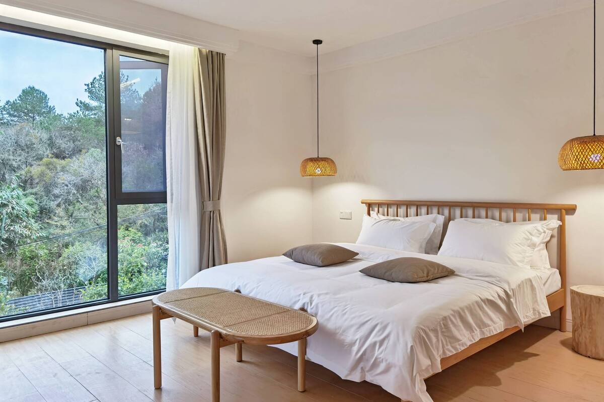 A bedroom with a large window, a bed with white bedding, two hanging lamps, and a wooden chair.