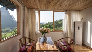 A wooden table with two chairs, a vase with flowers, and a view of greenery and mountains through large windows. - Riverside Retreat Hotel (Guilin)