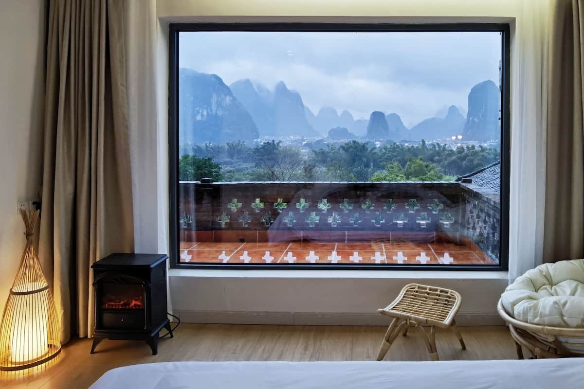 A room with a large window offering a view of mountains and greenery. A bed is positioned under the window, and there is a small table with a lamp beside it.