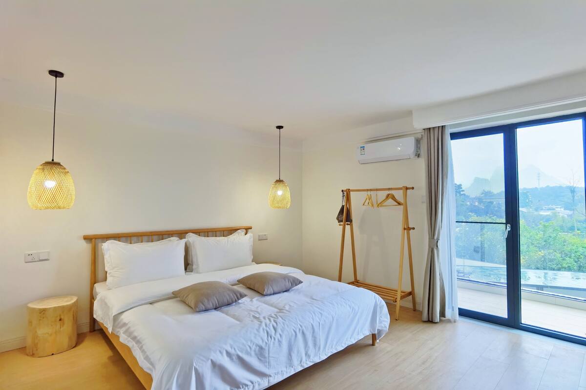 A bedroom with a large bed, two hanging lamps, a wooden bedside table, a wardrobe, and a balcony with a view.
