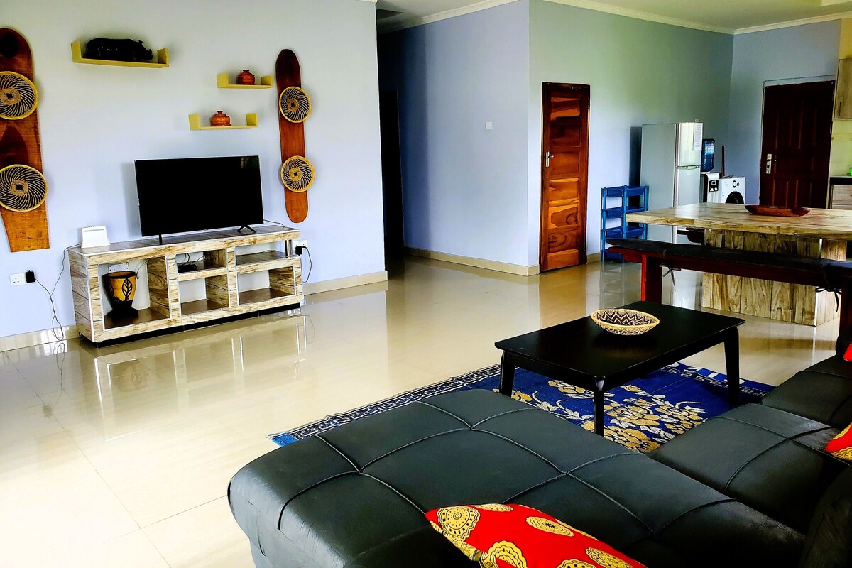 A living room with a black sofa, a TV on a wooden stand, a dining area with a wooden table, and a kitchen visible through an open door.