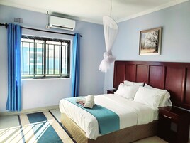 A hotel room with a bed, a nightstand, a window with curtains, an air conditioning unit, and a framed picture on the wall.