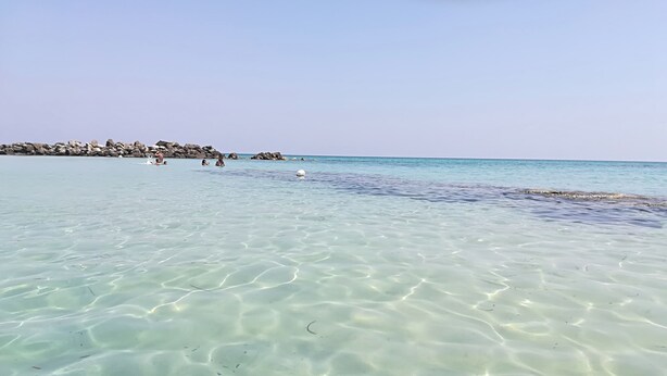 Una spiaggia assolata con acque basse, rocce sullo sfondo e alcune persone in lontananza.