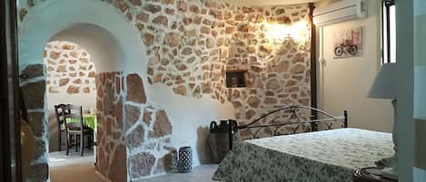 A bedroom with a stone wall, a bed with a floral bedspread, a nightstand, a lamp, and a small table with a chair.
