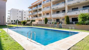 A swimming pool with a concrete deck, surrounded by a well-maintained lawn and a multi-story building with balconies.