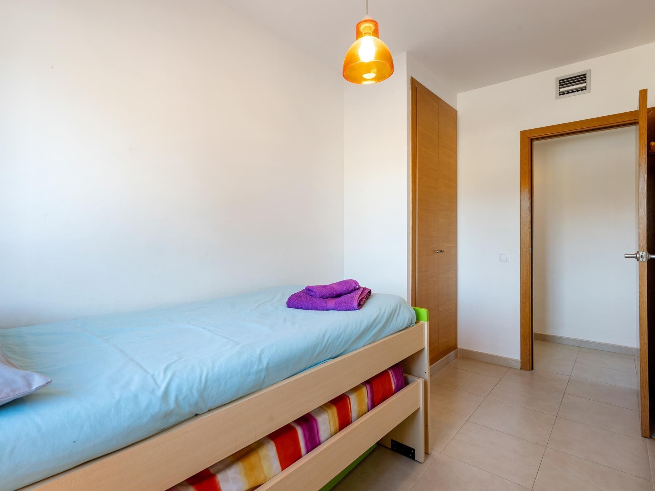 Un llit individual amb matalàs blau clar i tovallola morada, un armari de fusta i una porta que condueix a una altra habitació.