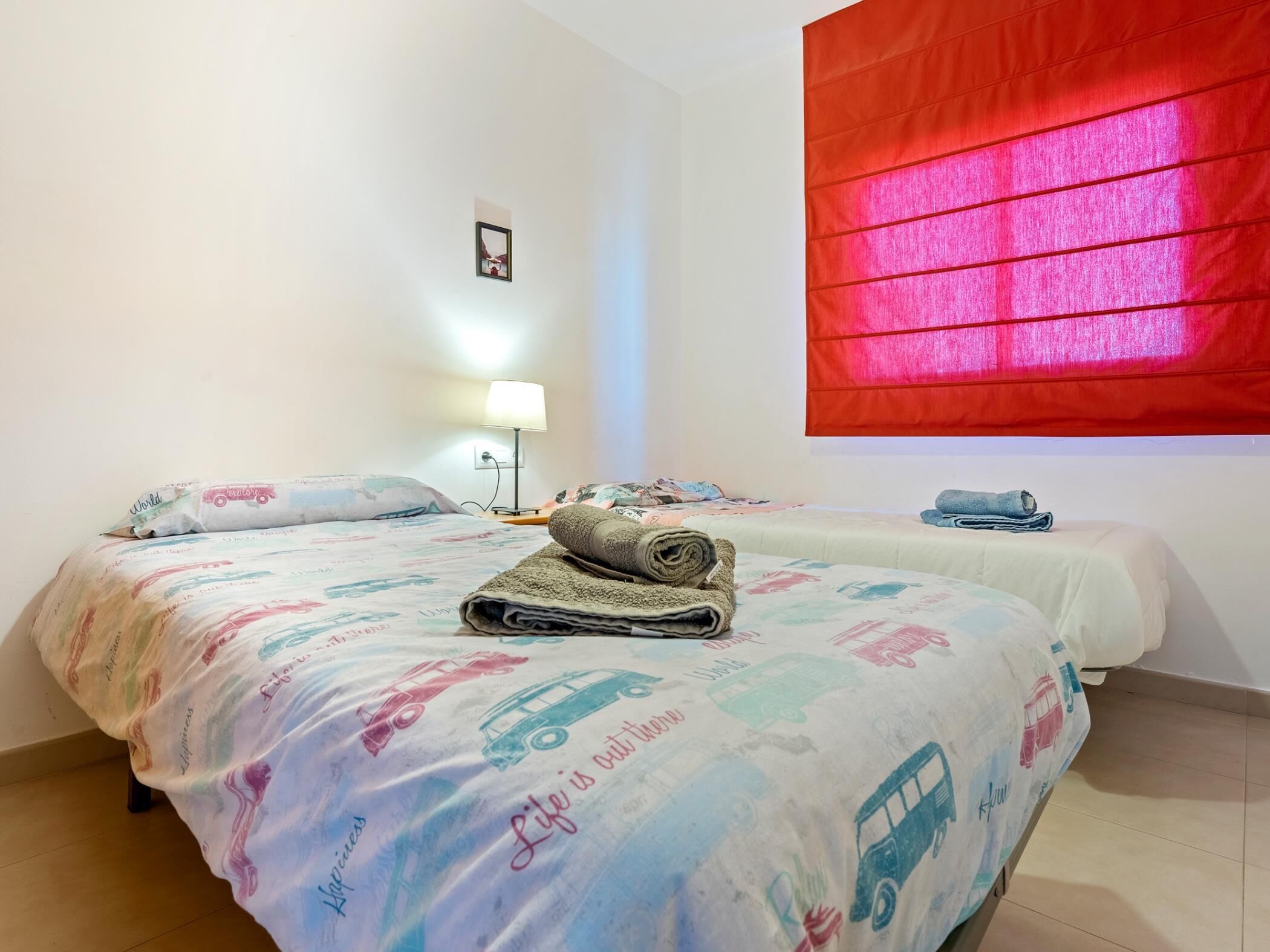 A hotel room with two beds, each with a colorful bedspread featuring vehicles and the phrase "Life is out." A red curtain is visible on the window, and a small table lamp is on the nightstand.