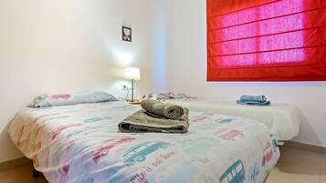 A hotel room with two beds, each with a colorful bedspread featuring vehicles and the phrase "Life is out." A red curtain is visible on the window, and a small table lamp is on the nightstand.