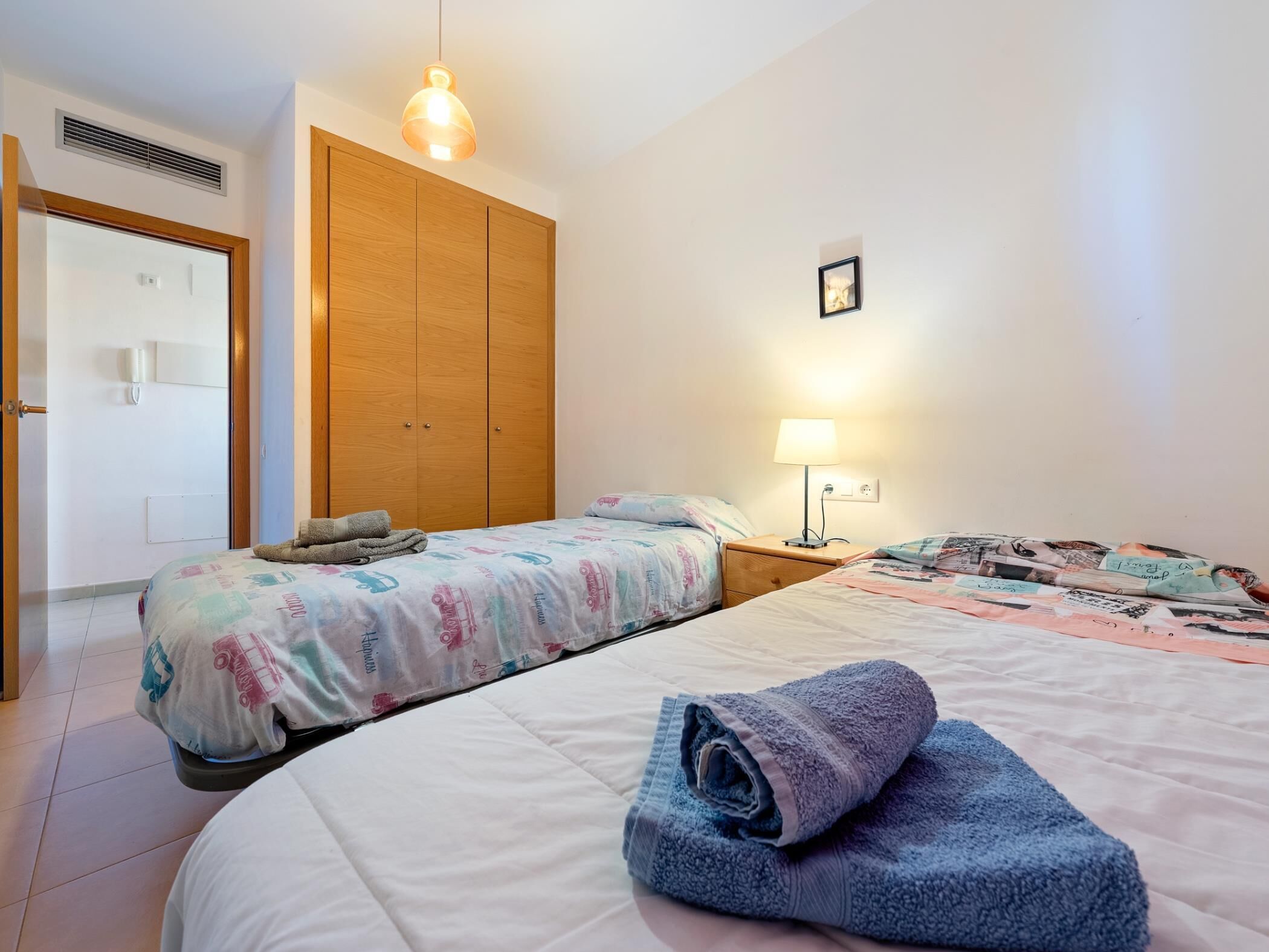 Une chambre avec deux lits, une armoire en bois, une table de chevet avec une lampe et un luminaire suspendu.