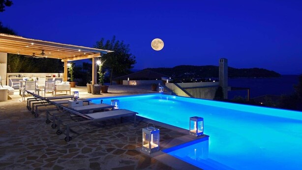 Une piscine entourée de chaises longues et d’une pergola, le tout agrémenté d’un sol en pierre et de plantes en pot, avec une vue sur la mer et la lune dans le ciel.