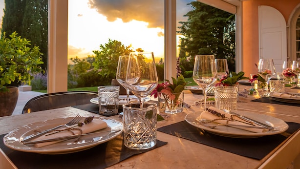 Una mesa preparada para una comida con cristalería transparente, platos y cubiertos, con vistas a árboles y un atardecer de fondo.