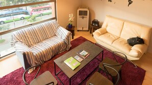 A modern living room with a striped sofa, a beige couch, a coffee table, and two chairs. There are hanging light fixtures, a world map on the wall, and a window with a view of a bus. - Seaview Capsule Hotel (Kota Kinabalu)