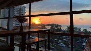 A room with a large window overlooking a sunset, a wooden table, and a hanging light fixture. - Seaview Capsule Hotel (Kota Kinabalu)