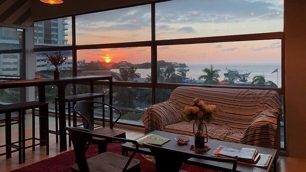 A modern living room with a large window, a sofa, a coffee table, and a dining area. - Seaview Capsule Hotel (Kota Kinabalu)