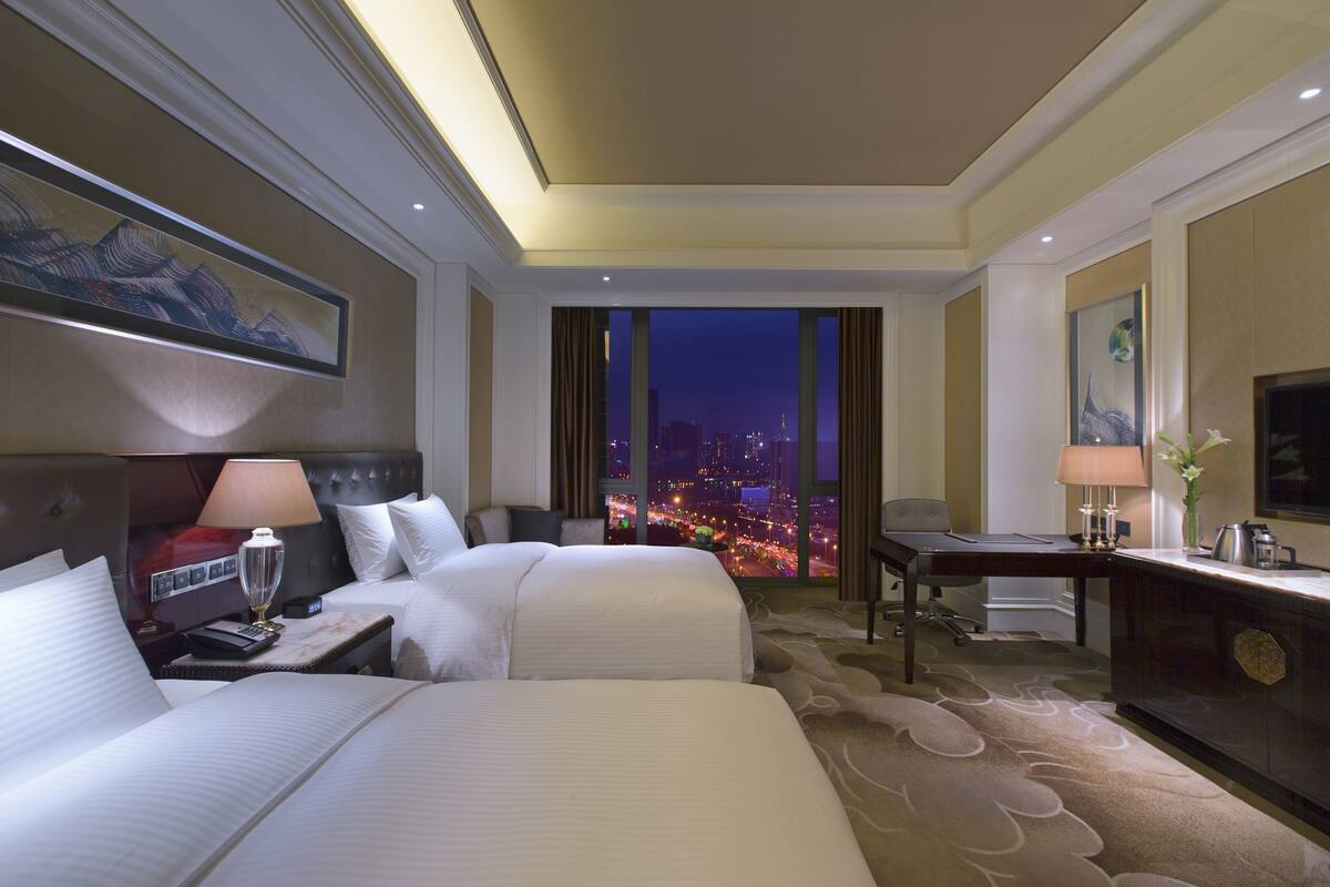 A hotel room with two beds, a desk, a chair, a TV, and a view of the city at night.