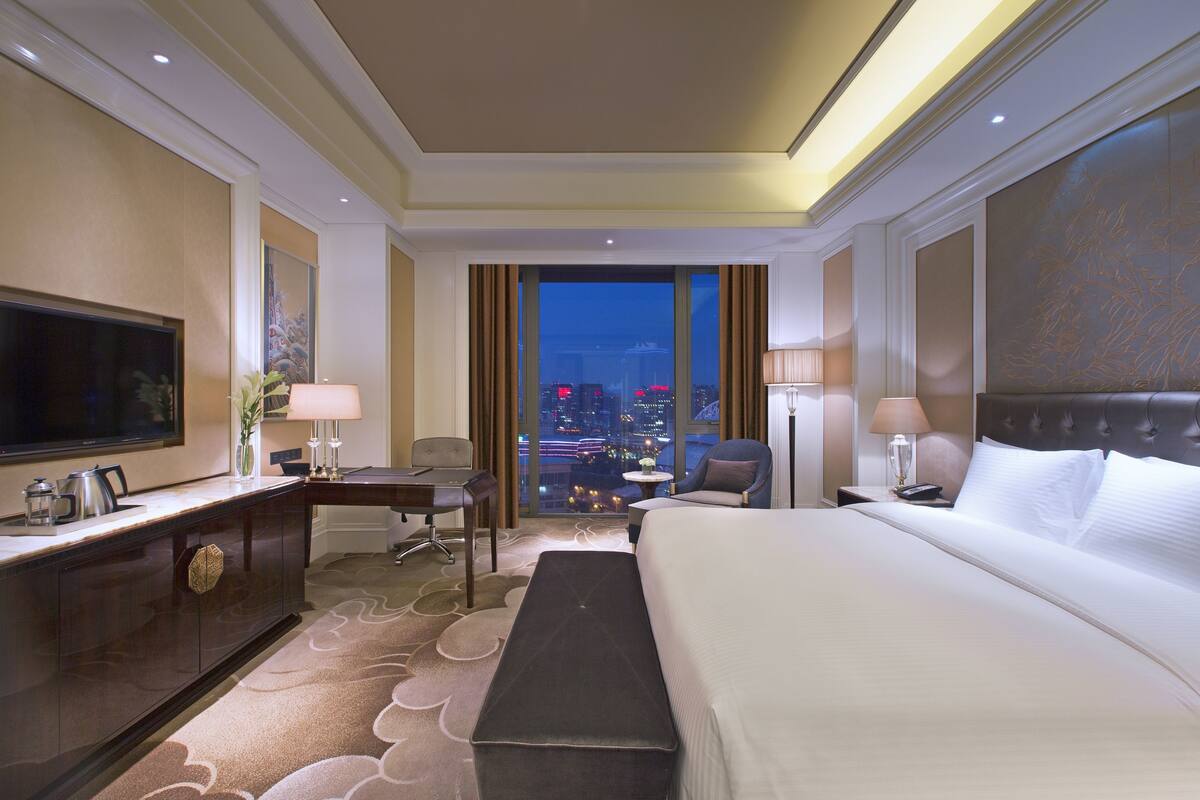 A hotel room with a large bed, a desk with a chair, a television, and a view of the cityscape.