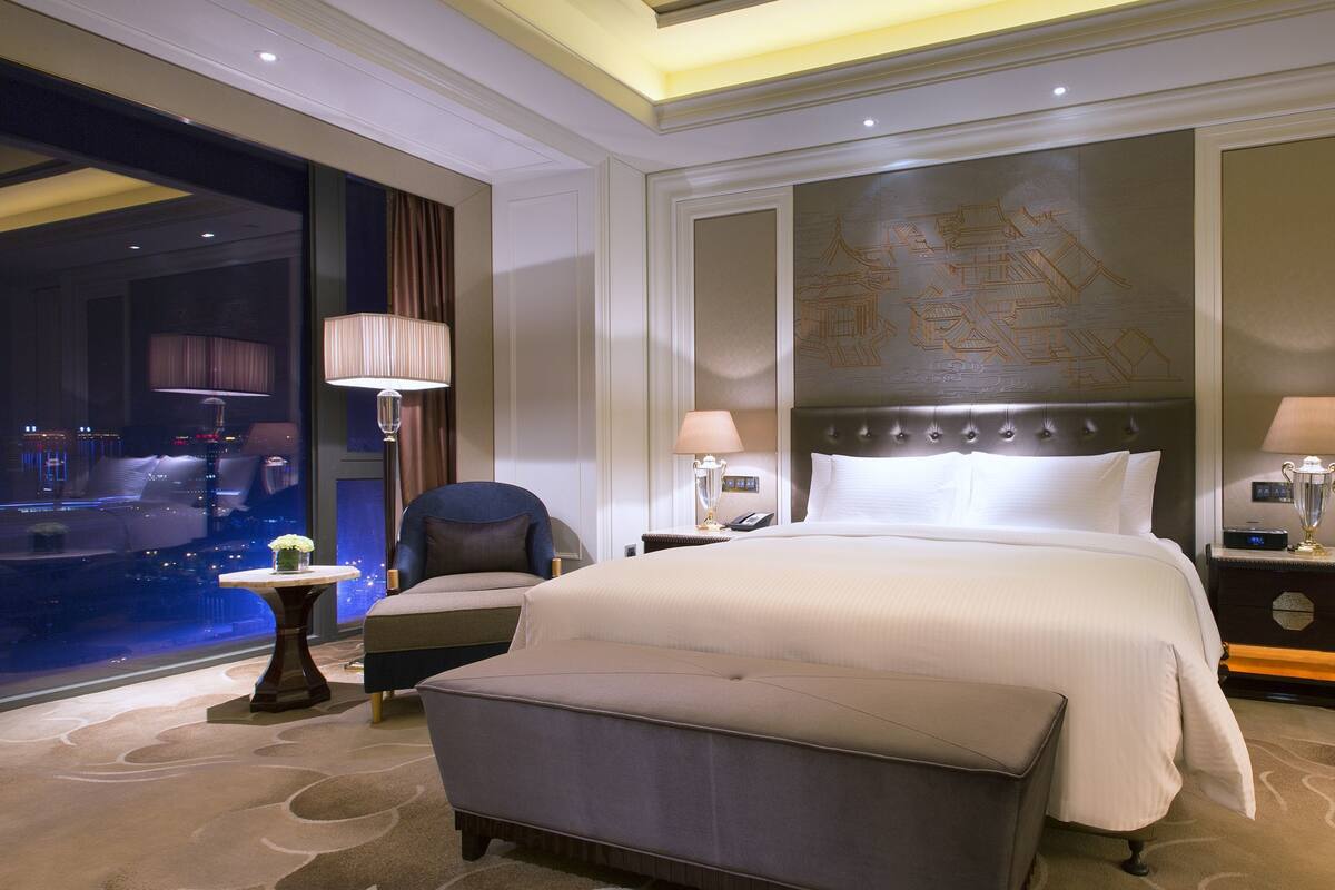 A hotel room with a large bed, two bedside tables, a chair, a small table with a vase, and a view of the city at night.