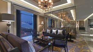 A spacious living room with a large sofa, a coffee table, and a chandelier. There is a view of the city through large windows.