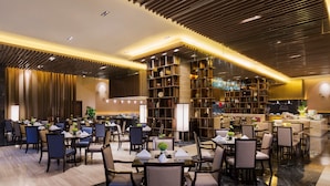 A modern dining area with multiple tables and chairs, a central decorative plant, and a large bookshelf. - Wanda Realm Bengbu (Bengbu)