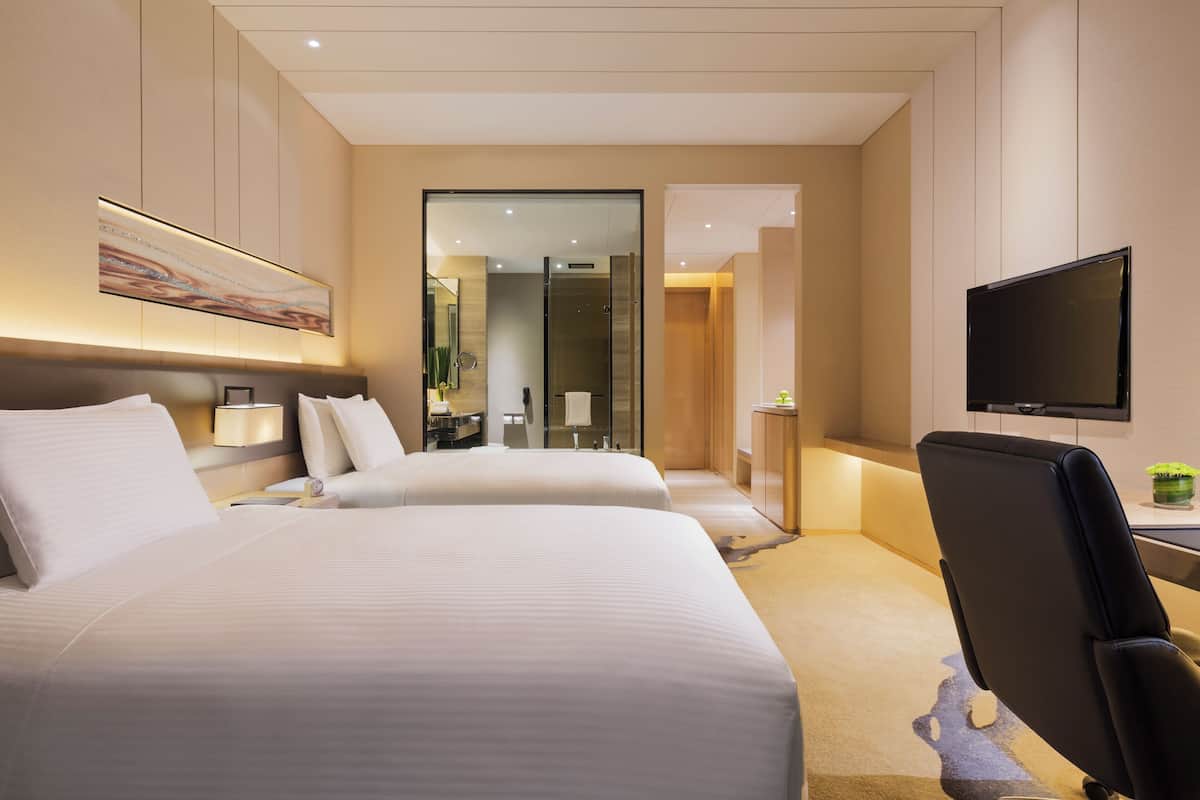 A hotel room with two beds, a TV, and a bathroom with a glass shower.