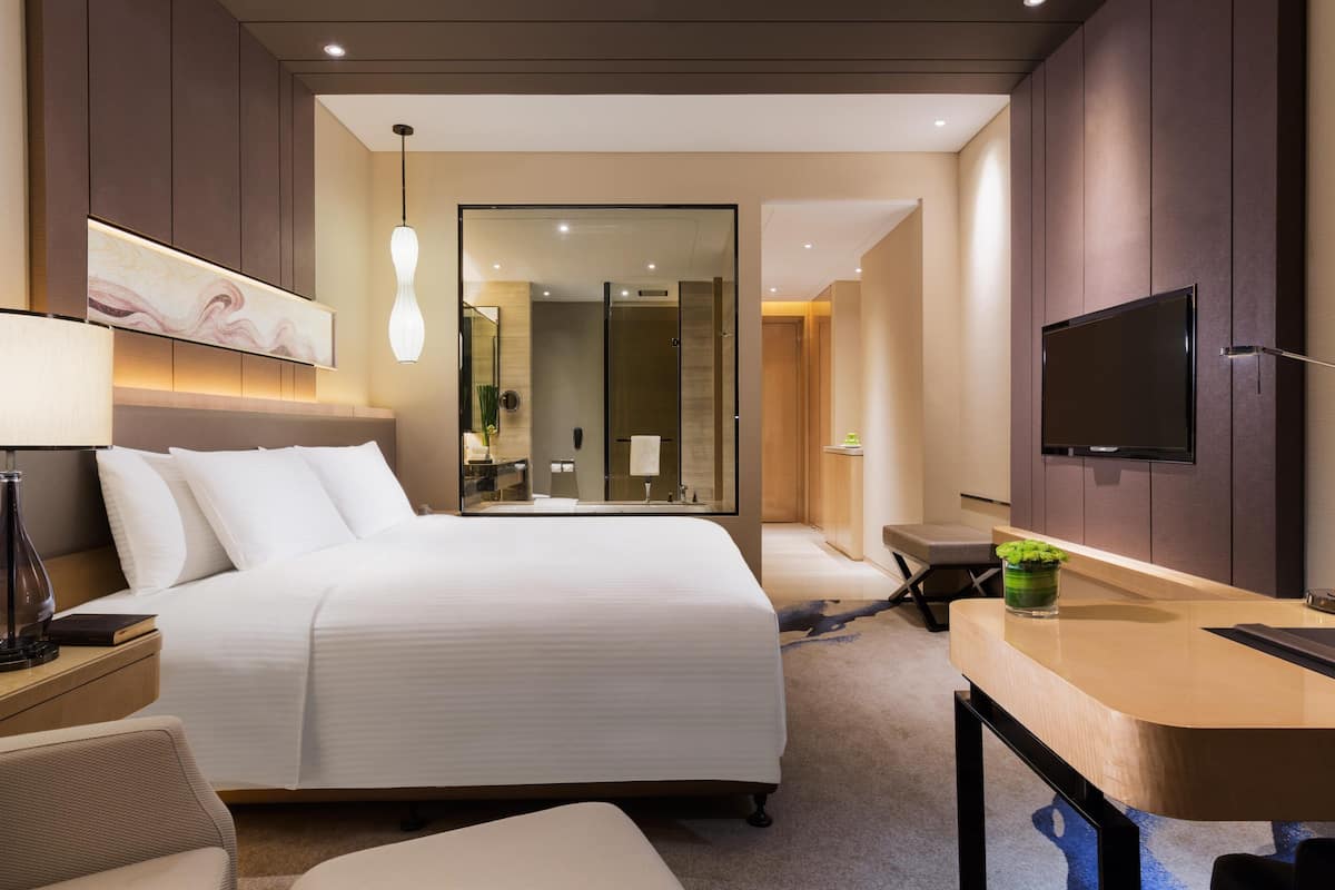 A modern hotel room with a large bed, a desk, a TV, and a bathroom visible through a glass door.