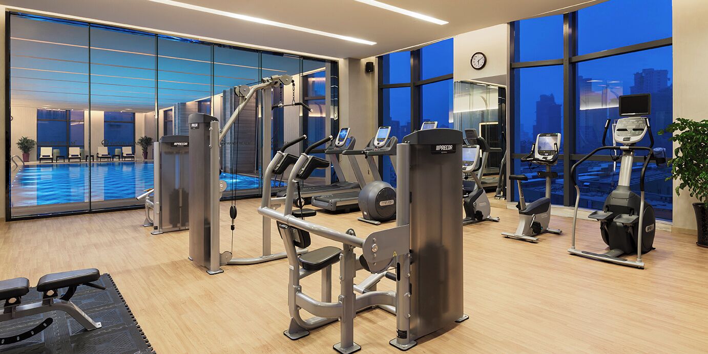 A modern gym with various exercise machines, a large window with a city view, and a pool area visible through the glass doors.