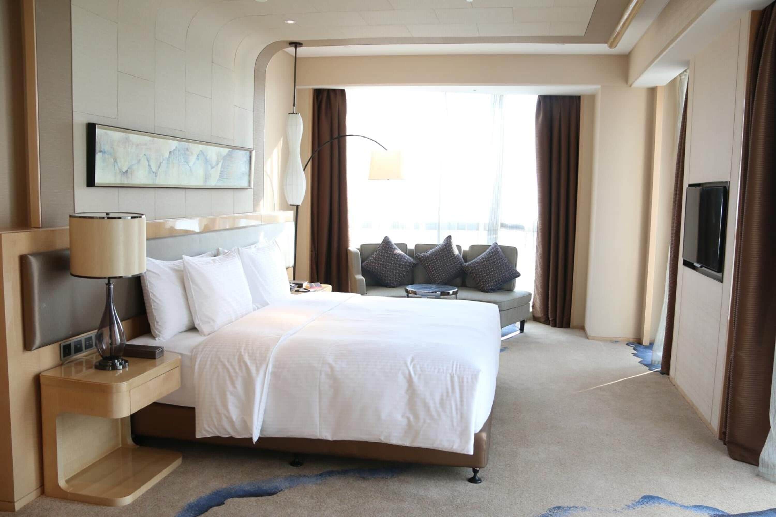Habitación de hotel con una cama grande, una mesita de noche con lámpara, un sofá con cojines, un televisor y un amplio ventanal con cortinas.