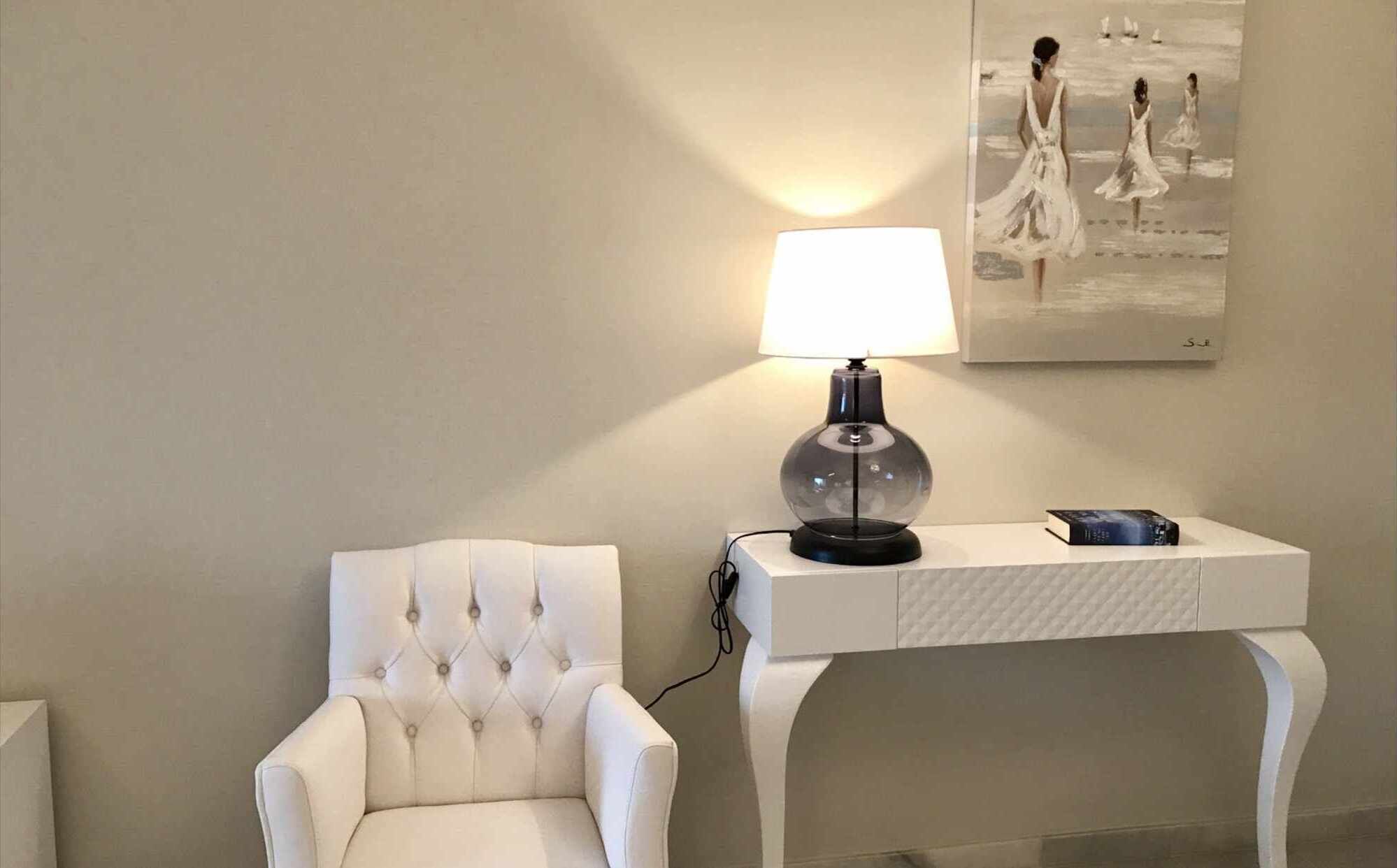 A white tufted armchair next to a white console table with a lamp and a book.