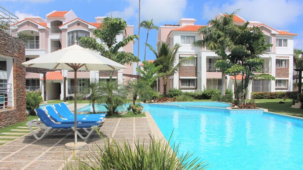 A swimming pool with lounge chairs and umbrellas, surrounded by palm trees and a building with a tiled roof. - CORTE SEA Luxury apartment in the heart of Bavaro (Punta Cana)